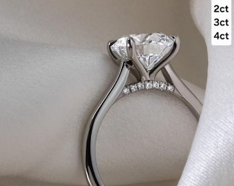 以下が含まれることがあります： 大きな透明なダイヤモンドが付いたシルバーの婚約指輪のクローズアップ。リングには、小さなアクセントストーンが付いた繊細なバンドがあります。右側のテキストは次のとおりです：1ct、2ct、3ct、4ct。
