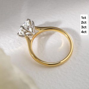 Op de afbeelding: Een gouden ring met een grote, heldere, gefacetteerde edelsteen in een zilverkleurige zetting. De ringband is van glad, gepolijst geelgoud. De tekst aan de rechterkant van de afbeelding toont de lijst: 1ct, 2ct, 3ct, 4ct.