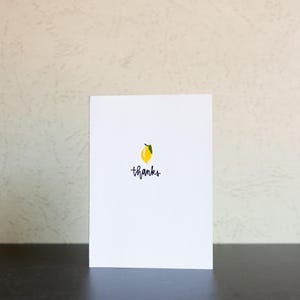 May include: A white card featuring a yellow lemon illustration with a green leaf and the word "thanks" in black script. The card is on a dark surface. The lemon is a bright, cheerful design.