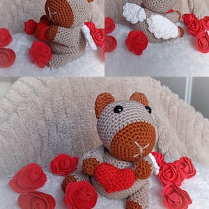 May include: A crocheted teddy bear with brown and tan yarn, holding a red heart. The bear has small white wings and is surrounded by red fabric roses. The bear is sitting on a white, fluffy surface.