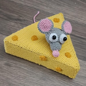 Puede incluir: Un ratón de ganchillo hecho a mano con cabeza gris, orejas y nariz rosas y ojos negros grandes se encuentra sobre una cuña de queso de ganchillo amarillo con agujeros de queso naranja. El ratón tiene un hilo rosa.