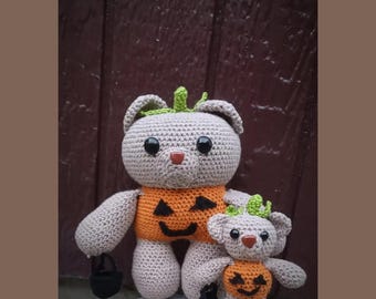 May include: Two crocheted teddy bears in light beige yarn, each wearing an orange pumpkin-themed sweater with a black jack-o'-lantern face. Both bears have black button eyes, brown noses, and small green stems on their heads. One bear holds a black bucket.