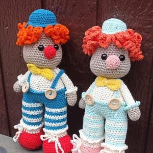 Op de afbeelding: Twee handgemaakte gehaakte clownpoppen. Elke pop heeft een grijze kop, oranje krullend haar, een rode neus en een gele vlinderdas. De ene pop draagt een blauw-wit gestreepte overall, de andere een lichtblauwe en wit gestreepte overall. Beide hebben rode schoenen.