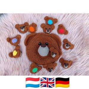 May include: A large brown crocheted bear face with black eyes and a light grey snout, surrounded by ten smaller brown crocheted bear appliques. Each small bear features a unique colourful crocheted accent on its forehead, including orange, blue, red, yellow, and green.
