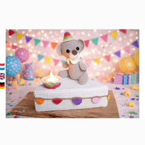 May include: A gray crocheted teddy bear wearing a colorful party hat sits on a white crocheted cake slice. The cake is decorated with a small rainbow-colored crocheted candle and a string of tiny, multi-colored crocheted triangular bunting.