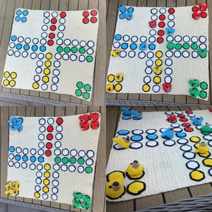 Op de afbeelding: Een crèmekleurig, gehaakt Ludo-bordspel met kleurrijke pionnen. Het bord heeft een kruisontwerp met blauwe, rode, groene en gele cirkels. De pionnen zijn gehaakte teddyberen in bijpassende kleuren.