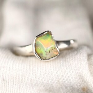 May include: A silver ring featuring a raw, multi-colored opal gemstone. The opal displays shades of green, yellow, and white. The ring has a simple, polished band.