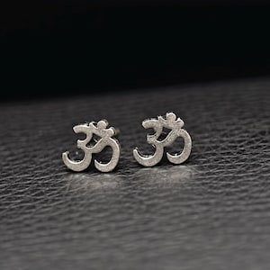 May include: A pair of silver-toned stud earrings featuring the Om symbol. The earrings have a textured, matte finish and a distinct cutout design that clearly displays the curves and lines of the sacred character in a small, compact size.