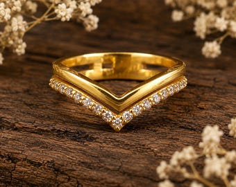 May include: A gold V-shaped ring featuring a polished band and a second band adorned with a row of small, clear, round-cut stones. The ring has a chevron design with a smooth finish on the upper section and a sparkling, stone-set lower section.