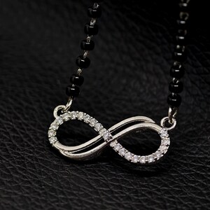 May include: A necklace featuring a silver-toned infinity symbol pendant encrusted with small clear stones. The pendant is attached to a chain composed of small, round, black beads that extend upwards from both sides of the infinity charm.