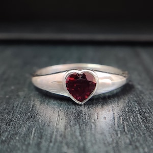 Könnte beinhalten: Ein silberner Ring mit einem herzförmigen, tiefroten Edelstein. Das Band des Rings ist glatt und poliert, wobei der Edelstein in einer Zargenfassung gefasst ist. Der Edelstein hat einen facettierten Schliff.