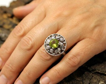 Può includere: Un anello in argento con una pietra preziosa rotonda e sfaccettata color verde lime al centro. La pietra è circondata da una cornice circolare decorata con dettagliati motivi a foglia in rilievo e piccole perline d'argento testurizzate.