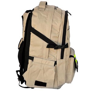 K&ouml;nnte beinhalten: Beigefarbener Rucksack mit schwarzen Riemen und Akzenten. Er verf&uuml;gt &uuml;ber mehrere Taschen, einen oberen Griff und ein Hauptfach mit Rei&szlig;verschluss. Der Rucksack hat ein gepolstertes R&uuml;ckenteil und verstellbare Schultergurte. Ein kleiner, reflektierender gelber Streifen ist sichtbar.