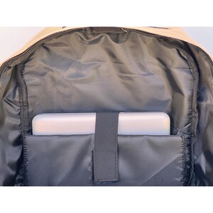 K&ouml;nnte beinhalten: Innenansicht eines beigen Rucksacks. Der Innenraum ist mit dunkelgrauem Stoff ausgekleidet und verf&uuml;gt &uuml;ber ein gepolstertes Fach, das einen wei&szlig;en Laptop aufnimmt. Ein schwarzer Gummizug sichert den Laptop. Der Rucksack ist offen und zeigt den Rei&szlig;verschluss.