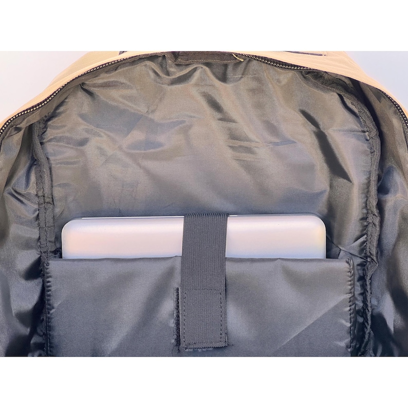 K&ouml;nnte beinhalten: Innenansicht eines beigen Rucksacks. Der Innenraum ist mit dunkelgrauem Stoff ausgekleidet und verf&uuml;gt &uuml;ber ein gepolstertes Fach, das einen wei&szlig;en Laptop aufnimmt. Ein schwarzer Gummizug sichert den Laptop. Der Rucksack ist offen und zeigt den Rei&szlig;verschluss.