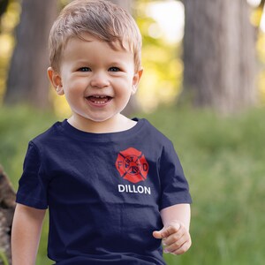 Happy toddler wearing a navy blue shirt with red Maltese cross on left chest and name underneath in white.