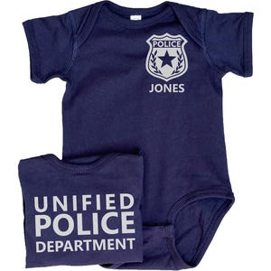 May include: A navy blue baby onesie with a white police badge and the name "JONES" printed on the front. The back of the onesie says "UNIFIED POLICE DEPARTMENT" in white.