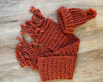 May include: A rust-colored knitted scarf with long fringe and a matching knitted hat with a pom-pom.