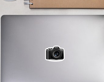 May include: A black camera sticker on a laptop. The sticker has the word "Camera" printed on it. A white computer mouse, a brown notebook, a pencil, a pair of glasses, and small potted plants are also visible.