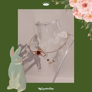 Peut inclure: Un collier ras de cou en dentelle blanche délicate avec une chaîne dorée et un pendentif en forme de cœur rouge avec des ailes dorées. Le pendentif a une pierre précieuse rouge au centre.