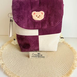 Könnte beinhalten: Ein Rucksack aus Cord mit einem Karomuster in Dunkellila und Creme. Die Frontklappe ist mit einem kleinen, gestickten beigen Bärengesicht-Aufnäher verziert. Ein rechteckiges Etikett auf der Vorderseite zeigt den Text "CreaTine HAND MADE".
