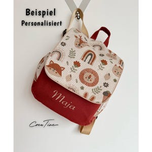 Könnte beinhalten: Ein kleiner Rucksack mit dunkelroter Basis und einer Klappe aus beigem Stoff mit Mustern aus Löwen, Füchsen, Rehen und Regenbögen. Der Name Maja ist in Weiß auf den roten Bereich gestickt. Der Text Beispiel Personalisiert ist zu sehen.
