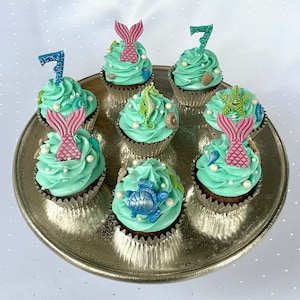 Könnte beinhalten: Sieben Cupcakes zum Thema Meerjungfrau, verziert mit rosa und blauen Fondant-Meerjungfrauen, Muscheln und türkisfarbenem Frosting. Die Cupcakes sind auf einer silbernen Platte angeordnet.