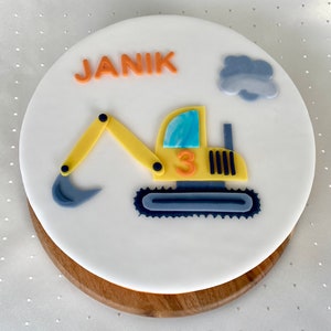 Könnte beinhalten: Ein weißer Kuchen, verziert mit einem gelben Bagger, einer blauen Wolke und dem Namen "JANIK" in orangefarbenen Buchstaben. Der Bagger hat eine blaue Schaufel und die Nummer "3" an der Seite.
