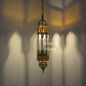 May include: A hanging pendant lamp with a hexagonal metal frame in an antique brass finish and amber-colored glass panels. The lamp features intricate cut-out metal detailing at the top and bottom, suspended by a chain. Text reads World of Lamp.