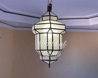 May include: A white glass and black metal Moroccan pendant light fixture hangs from a chain. The light fixture has a geometric design and is lit from within.