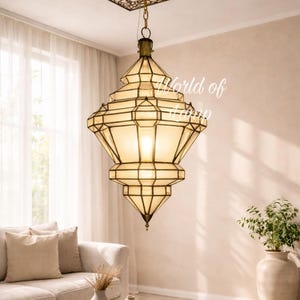 May include: A geometric pendant light fixture with a warm glow hangs from the ceiling. The light has a multi-tiered design with a brass finish and cream-colored glass panels. The ceiling features a decorative, ornate metal panel. The room is decorated in neutral tones.