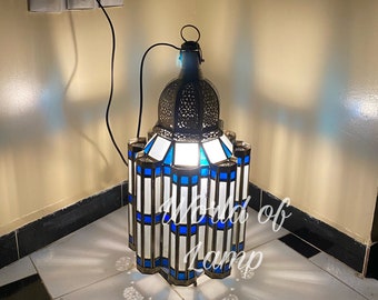 May include: A blue and white stained glass Moroccan lantern with a metal frame. The lantern is lit and casting a warm glow. The lantern is plugged into an electrical outlet.