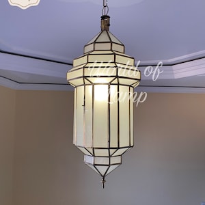 Pode incluir: Uma grande lâmpada de pendente de latão adornada com um abajur de vidro branco. A luz está pendurada em uma corrente e tem um design geométrico.