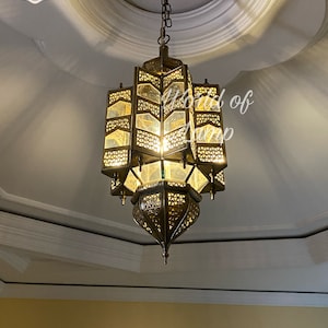 May include: A gold metal Moroccan style chandelier with intricate geometric patterns and clear glass panels. The chandelier is hanging from a white ceiling.
