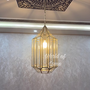 May include: A hanging gold-colored lantern with a geometric design, suspended from a chain. The lantern has a glass enclosure and a lit bulb inside. The ceiling features a decorative pattern. The text "World of Lamp" is visible.