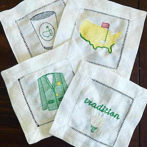 May include: Four white linen napkins with green embroidery. One napkin features a golf cup, another a green jacket, another a golf ball with the word "tradition", and the last a map of the United States with a flag.