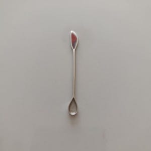 May include: A slender, silver-toned metal tool featuring a small, teardrop-shaped scoop at one end and a flat, curved, petal-like blade at the opposite end. The long, thin handle connects both ends of this dual-purpose implement.
