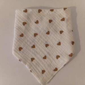 May include: White muslin baby bib with a pattern of brown hearts. The bib is triangular in shape and has a soft, textured appearance. The heart pattern is evenly spaced across the fabric.