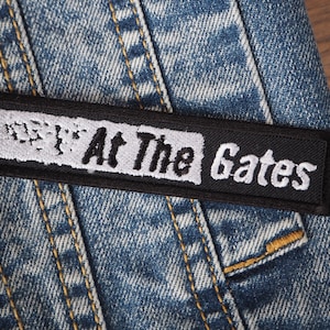 Puede incluir: Parche bordado en blanco y negro sobre una chaqueta vaquera azul. El parche dice "At The Gates".