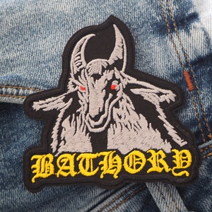 Puede incluir: Parche bordado en blanco y negro con una cabeza de cabra con ojos rojos y la palabra "BATHORY" en letras amarillas.