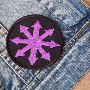 Puede incluir: Un parche redondo bordado en negro con un diseño morado de estrella de ocho puntas con flechas que apuntan hacia afuera. El parche está colocado sobre una chaqueta vaquera, con costuras visibles y un botón metálico.