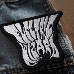 May include: Black and white embroidered patch with the text "WILD HEARTS" in a stylized font. The patch is sewn onto a denim jacket. The patch has a black border and is a unique accessory.