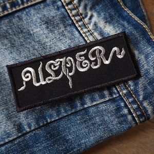 Puede incluir: Parche bordado negro con la palabra "USPER" en letras blancas en cursiva. El parche está cosido en una chaqueta vaquera azul.