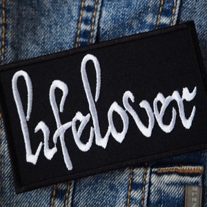 May include: Black fabric patch with white embroidered text that reads "Lifelover".