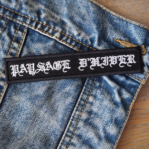 Puede incluir: Un parche negro con texto bordado en blanco que dice "PAY SAGE D'LIVER". El parche está cosido en una chaqueta vaquera azul.