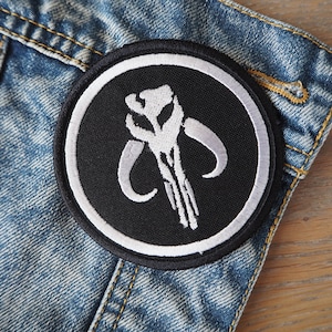 Può includere: Una toppa ricamata in bianco e nero con il simbolo mandaloriano su uno sfondo in denim.
