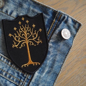 May include: A black embroidered patch with a gold tree and stars on a denim jacket.