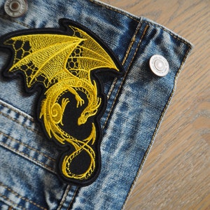 May include: A yellow embroidered dragon patch with black detailing. The patch is sewn onto a blue denim jacket.