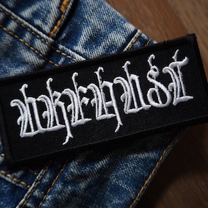 Puede incluir: Parche bordado negro con la palabra "Infest" en letras blancas sobre un fondo de mezclilla.