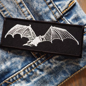 Puede incluir: Un parche bordado negro con un diseño de murciélago blanco. El parche está cosido en una chaqueta vaquera azul.